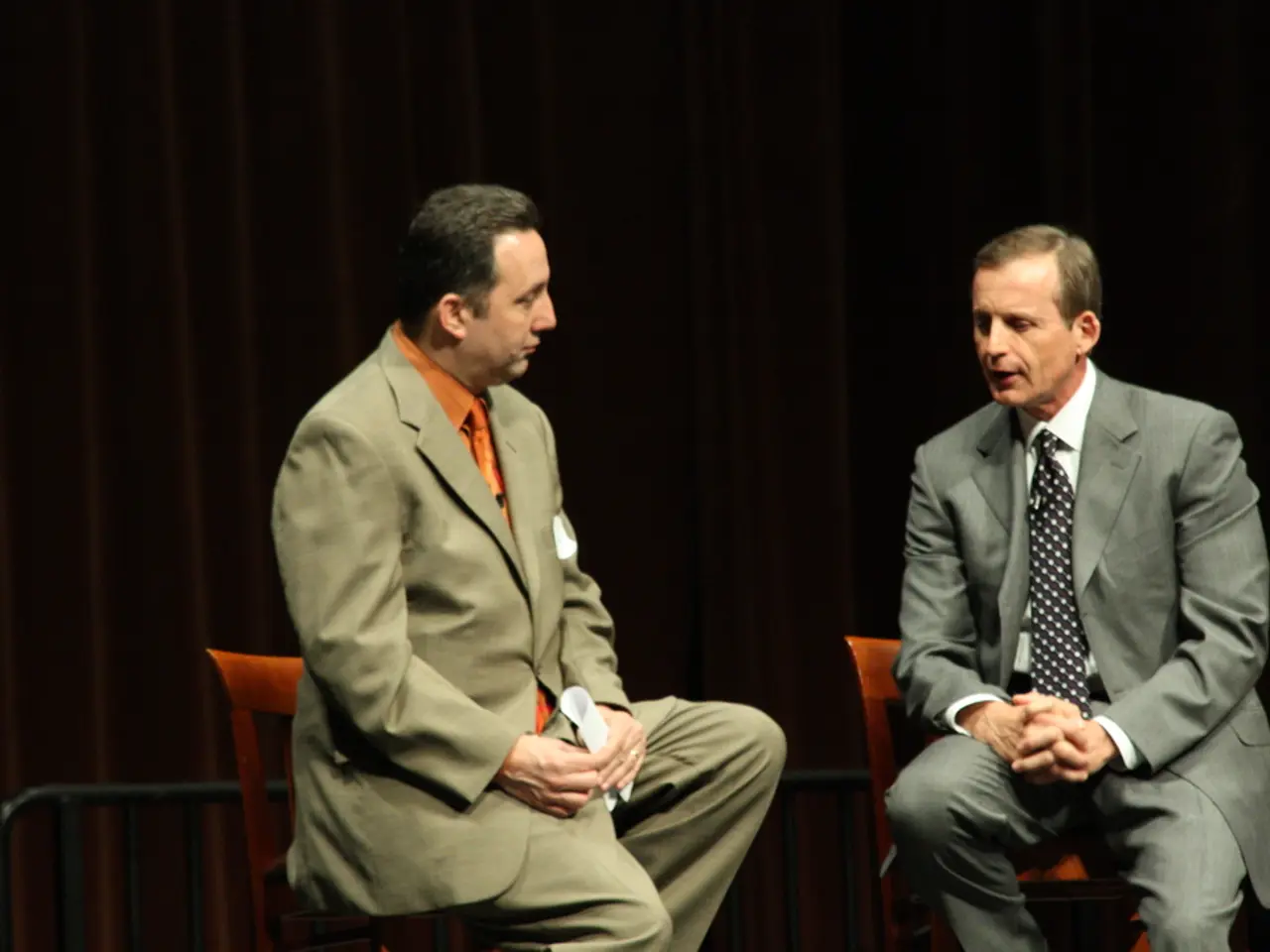 In this picture, there are two men sitting on the chairs. Both of them are wearing blazers,...