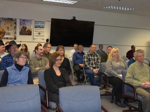 There are many people sitting in the chairs in this room. There are men and women in this group. In...