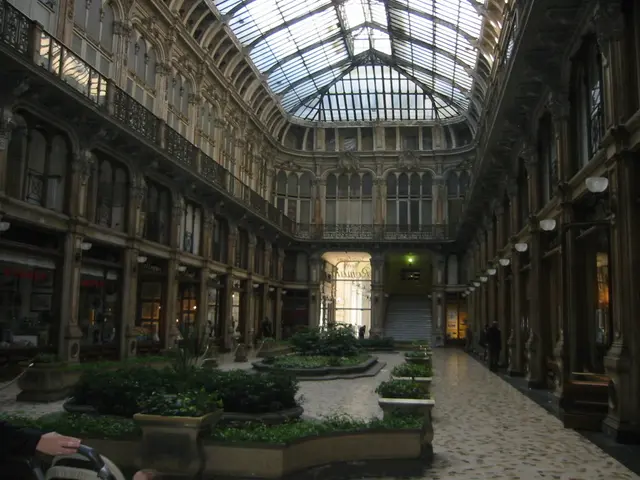 In this image we can see a building, electric lights, poles, name boards and houseplants.