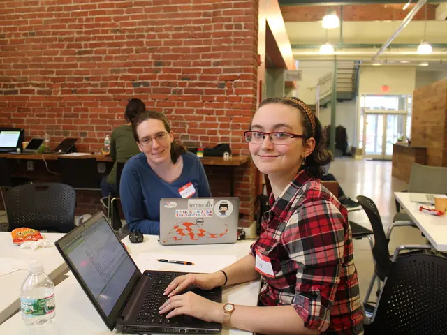 In this picture there are two woman sat on chair in front of table,there are two laptops in front...