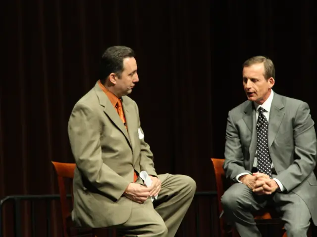 In this picture, there are two men sitting on the chairs. Both of them are wearing blazers,...